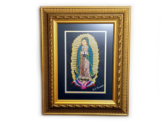 A framed religious art print of Our Lady of Guadalupe, featuring a gold ornate frame, a black mat, and a central illustration with gold rays, blue cloak, and pink doves, hanging on a light gray wall.