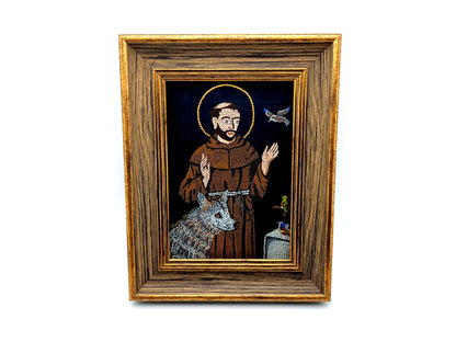 A framed religious icon depicting Saint Francis of Assisi with a brown robe, gold halo, and a silver wolf on a dark background, enclosed in a rustic brown wooden frame with gold trim.