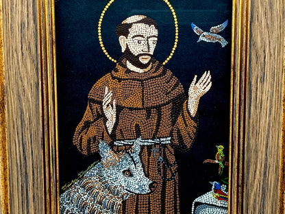 A framed mosaic religious artwork depicting Saint Francis of Assisi in a brown robe with a gold halo, alongside a wolf, dove, and small birds, set within a wood-grain frame.